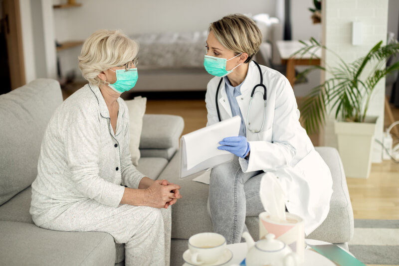 Female doctor and senior woman with protective face masks talking about medical data during a home visit